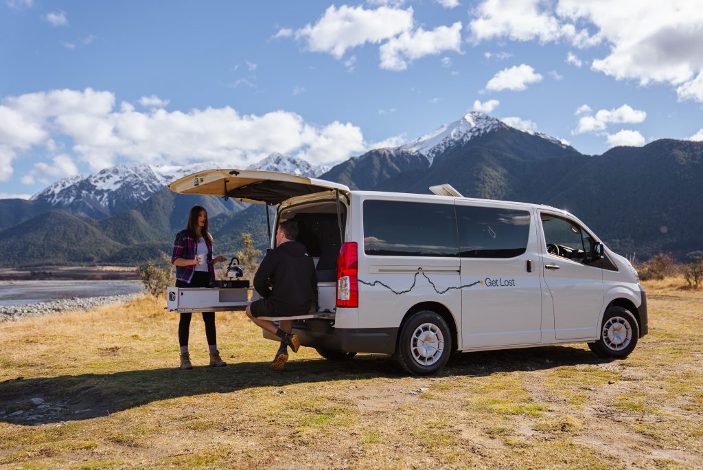 Couple at the open rear of a Get Lost campervan with a slide-out stove, parked in front of snow-capped mountains