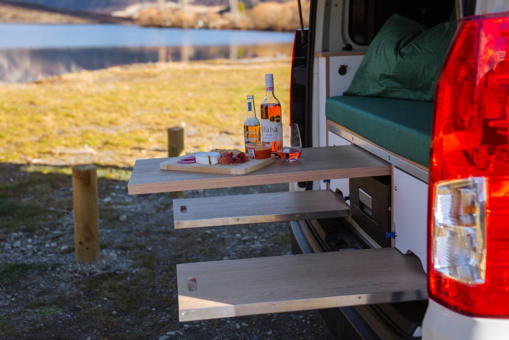 Rear of a Get Lost campervan with three slide-out wooden tables set for a picnic beside a New Zealand lake