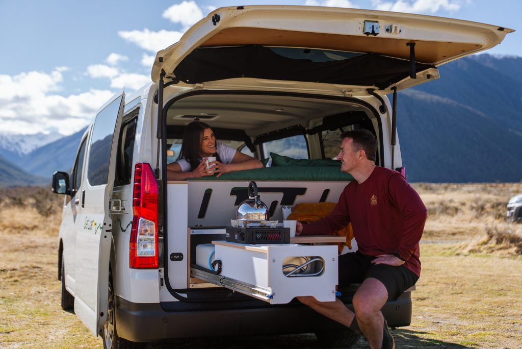 Couple relaxing in a Get Lost campervan with the bed and pull-out kitchen open in a mountain landscape
